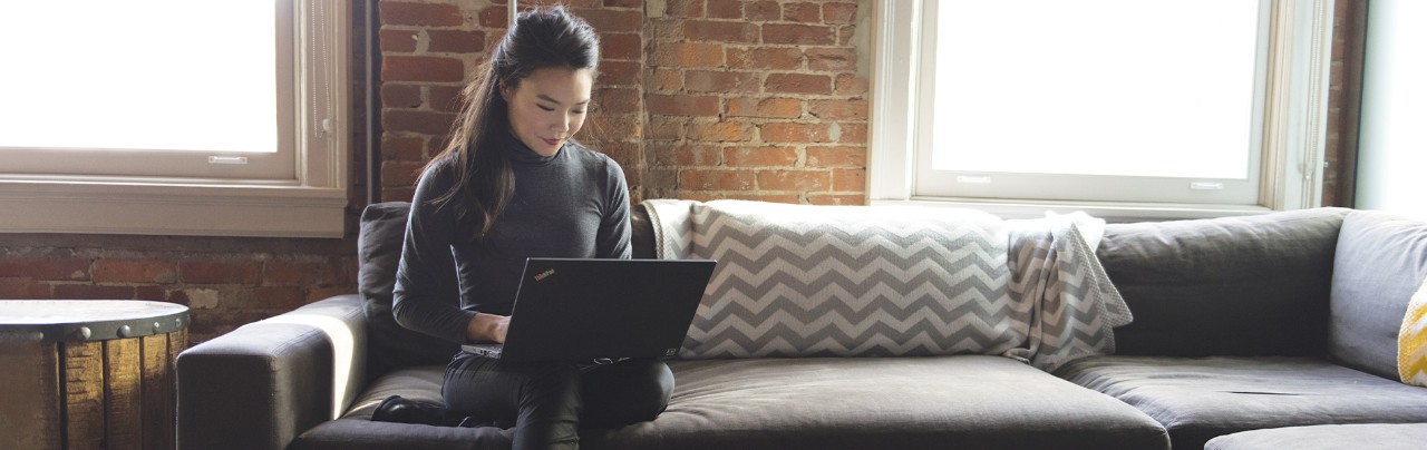 Woman sitting on a gray couch with a black laptop on her lap.